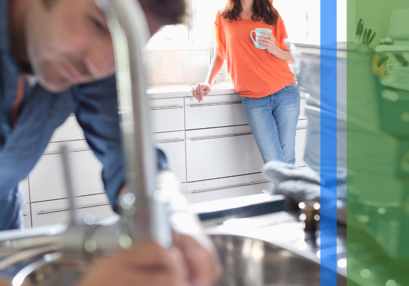 A plumber working on a sink with a woman watching in the background.