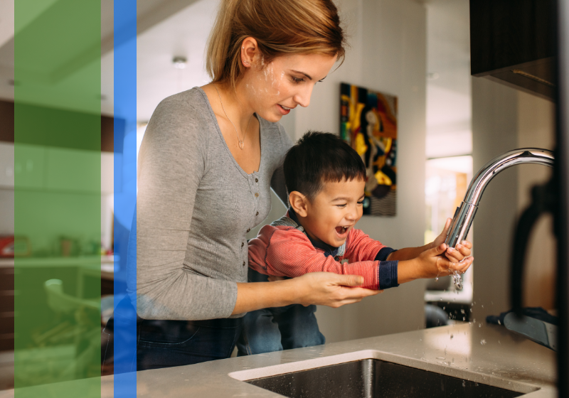 A woman holding up a young boy to a kitchen sink as he washes his hands.