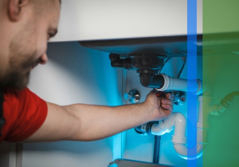 A plumber in a kitchen is working on a pipe under a counter.