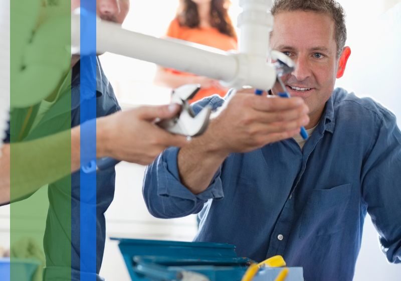 Two plumbers working on a pipe with a woman watching in the background
